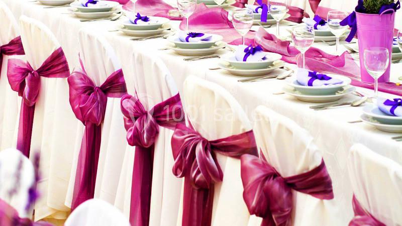 Chaises blanches décorées de nœuds roses alignées autour d'une table dressée avec vaisselle et serviettes violettes.