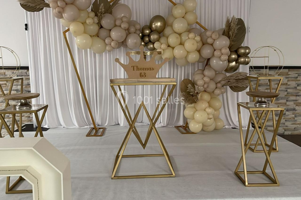 Décoration de fête avec arche de ballons beige et doré, support central en métal doré et panneaux blancs en arrière-plan.