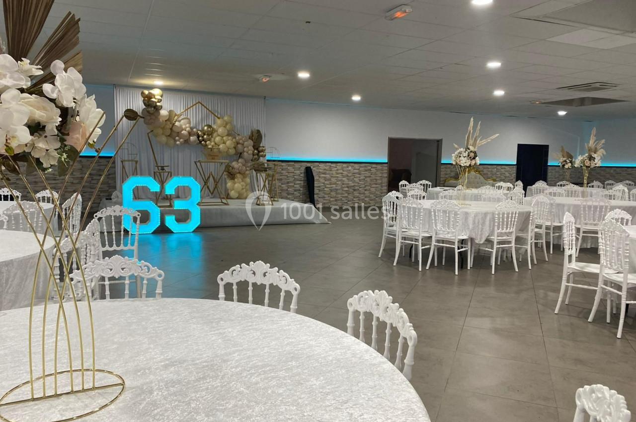 Salle de réception décorée avec des tables rondes, des chaises blanches et une scène ornée de ballons et chiffres lumineux.