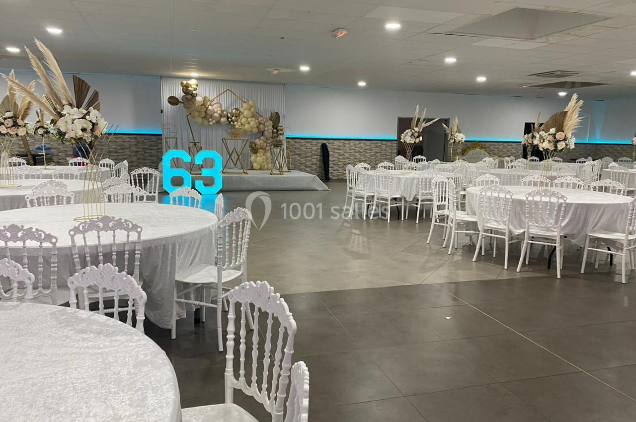 Salle de réception décorée avec des tables rondes couvertes de nappes blanches et une scène ornée de ballons et fleurs.
