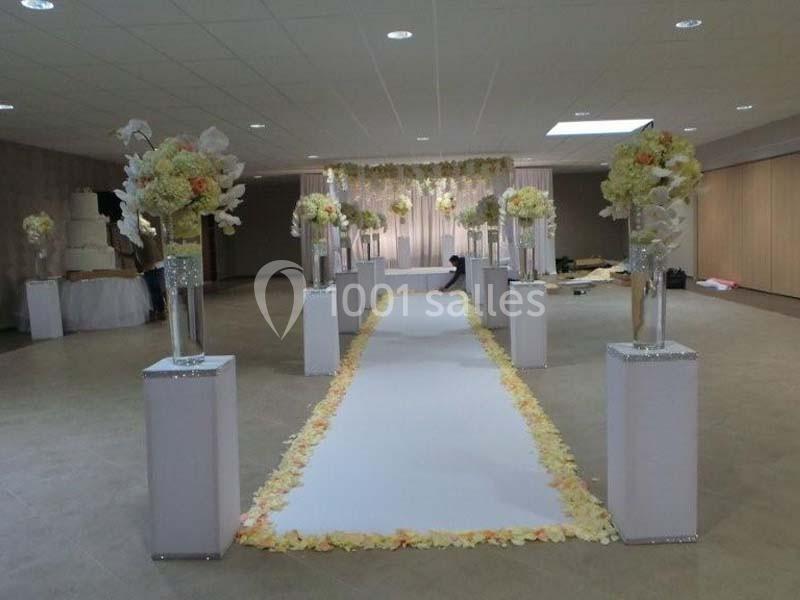 Allée de mariage décorée de pétales de fleurs et de colonnes ornées de bouquets dans une salle de réception.