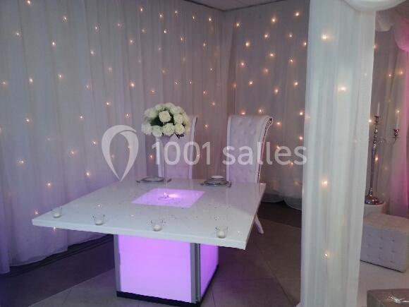 Table décorée avec des bougies, éclairage violet et chaises blanches, entourée de rideaux lumineux.