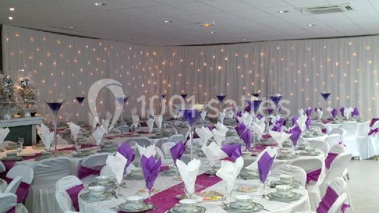 Salle de réception décorée avec des nappes blanches, des serviettes violettes pliées et des guirlandes lumineuses en fond.