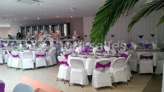 Salle de réception décorée avec des tables rondes, nappes blanches et chaises ornées de nœuds violets.