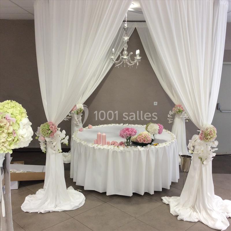 Table ronde décorée de fleurs roses et blanches, entourée de rideaux blancs et éclairée par un lustre.