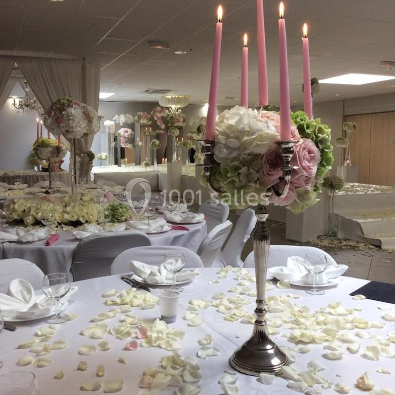 Centre de table avec chandelier rose, fleurs blanches et roses, sur une table décorée de pétales dans une salle de réception.
