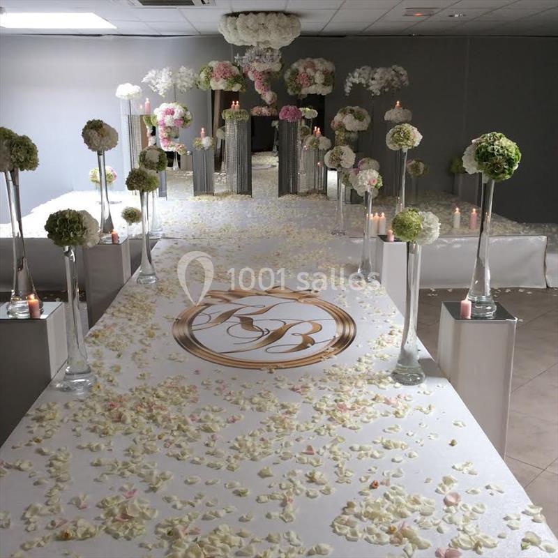 Allée de mariage décorée de pétales de fleurs, colonnes ornées de bouquets et logo central sur le tapis blanc.