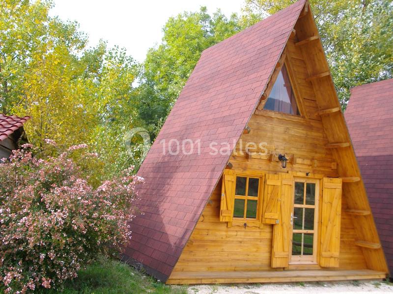 Chalet en bois en forme de A avec toit rouge, entouré de végétation et d'arbustes fleuris.