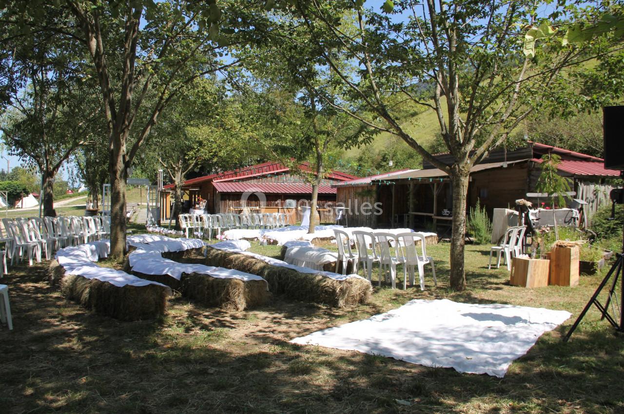 Chaises blanches et bottes de foin disposées en extérieur sous des arbres pour une cérémonie ou un événement.