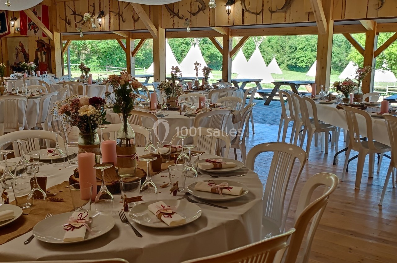 Salle décorée pour un événement avec tables rondes dressées, chaises blanches et vue sur un extérieur verdoyant avec tipis.