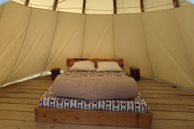 Tente tipi beige installée dans un champ verdoyant sous un ciel bleu, avec une ouverture laissant entrevoir l'intérieur.