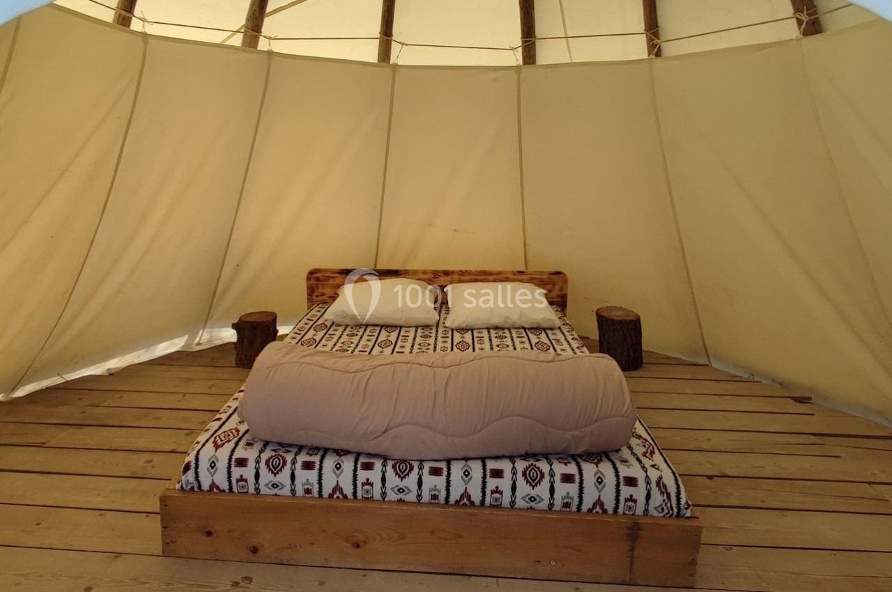 Lit double avec couverture beige et coussins blancs, installé dans une tente avec structure en bois et toile beige.