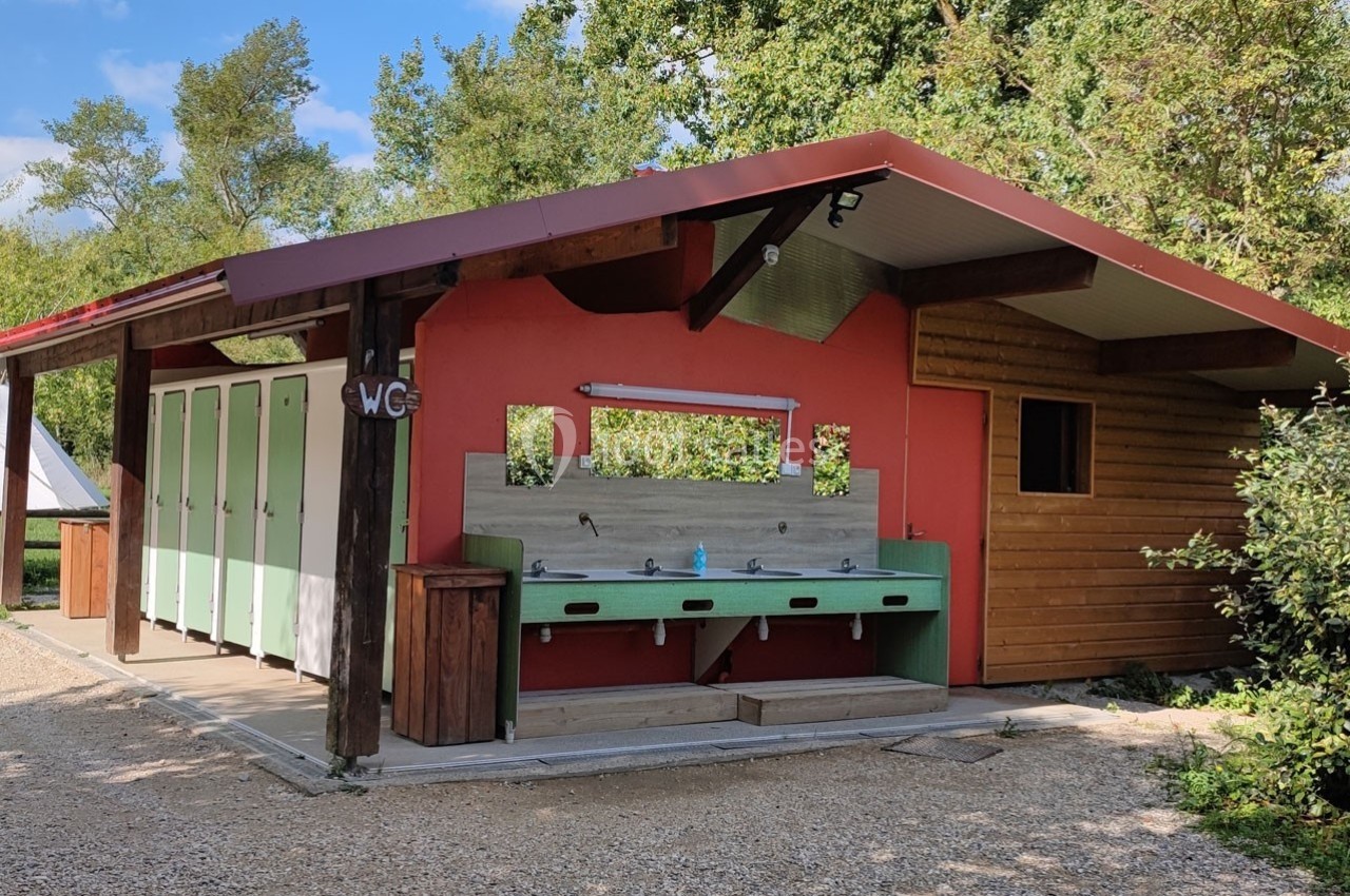 Bâtiment sanitaire avec cabines de toilettes, lavabos extérieurs et structure en bois dans un environnement arboré.