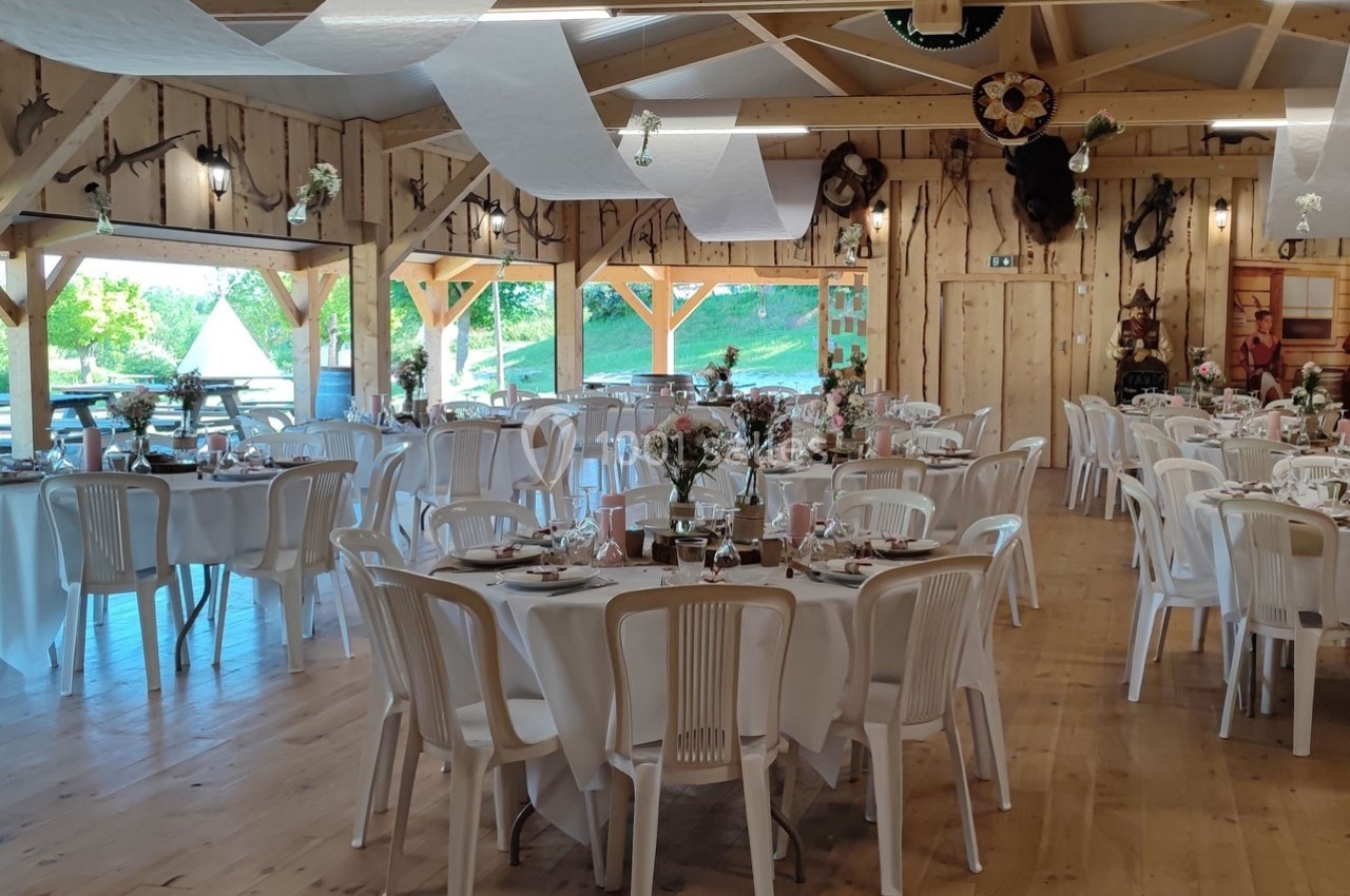Salle de réception en bois décorée pour un événement, avec tables rondes dressées et chaises blanches.