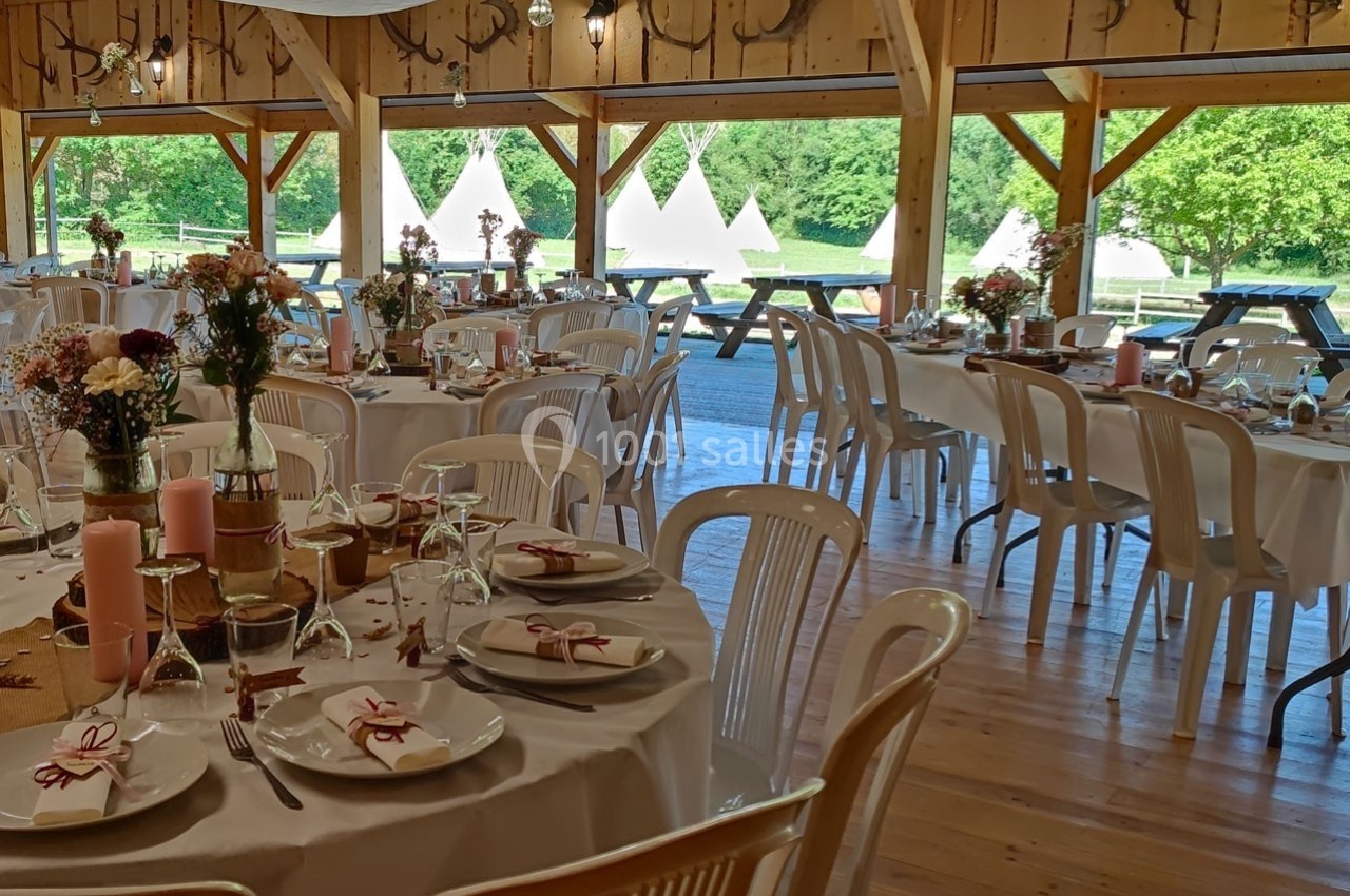 Salle de réception décorée pour un événement, avec des tables rondes dressées et vue sur un extérieur verdoyant.