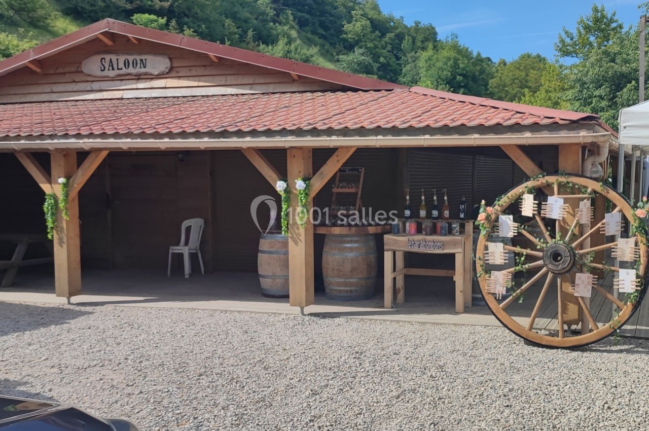 Façade d'un saloon en bois avec des tonneaux, une roue décorative et un stand de boissons devant.