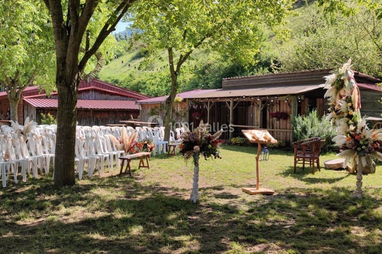 Chaises blanches alignées en extérieur sous des arbres, avec un pupitre et des décorations florales devant une grange en…