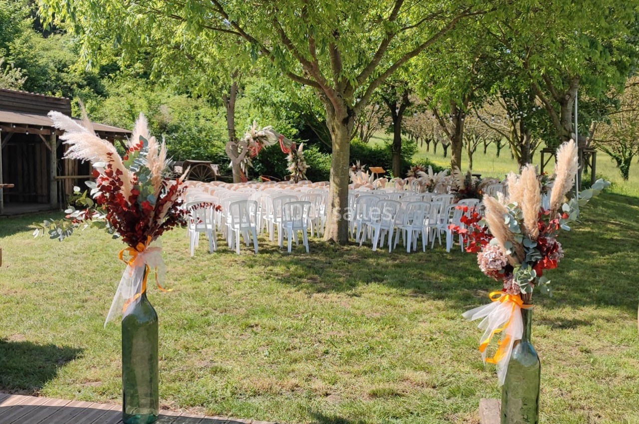 Chaises blanches disposées en extérieur sous des arbres, décorées avec des bouquets de fleurs et de pampas.