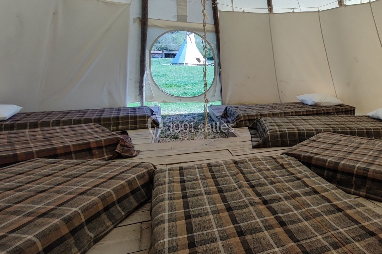 Intérieur d'un tipi avec plusieurs lits simples alignés, couvertures à carreaux, et vue sur un autre tipi à l'extérieur.