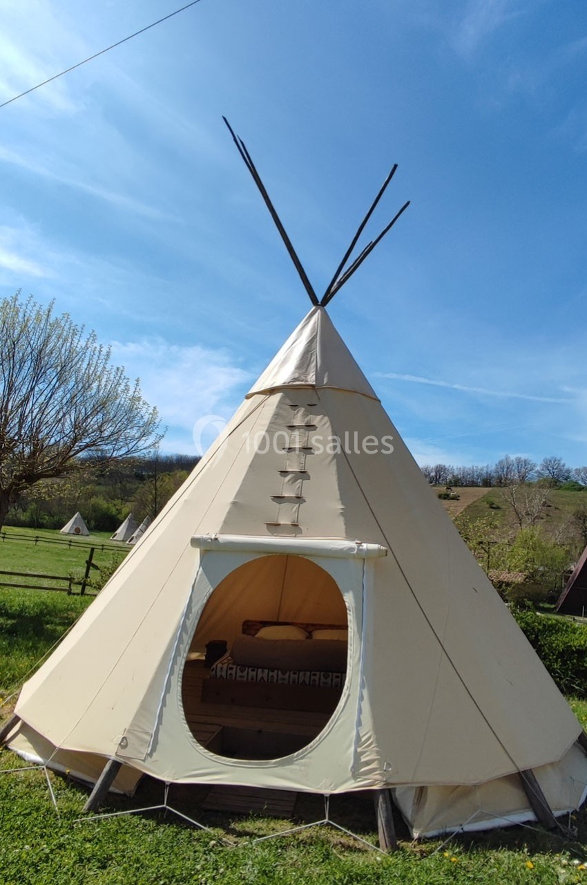 Tente tipi beige installée dans un champ verdoyant sous un ciel bleu, avec une ouverture laissant entrevoir l'intérieur.