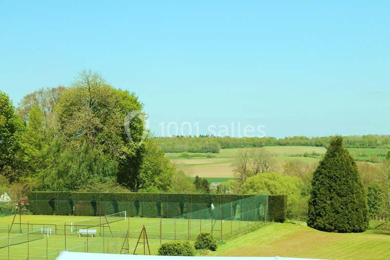 Vue sur un terrain de tennis entouré de haies, avec des champs et des arbres en arrière-plan sous un ciel dégagé.