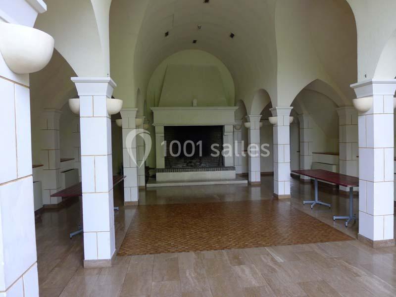 Salle voûtée avec cheminée centrale, colonnes blanches et tables pliantes le long des murs.