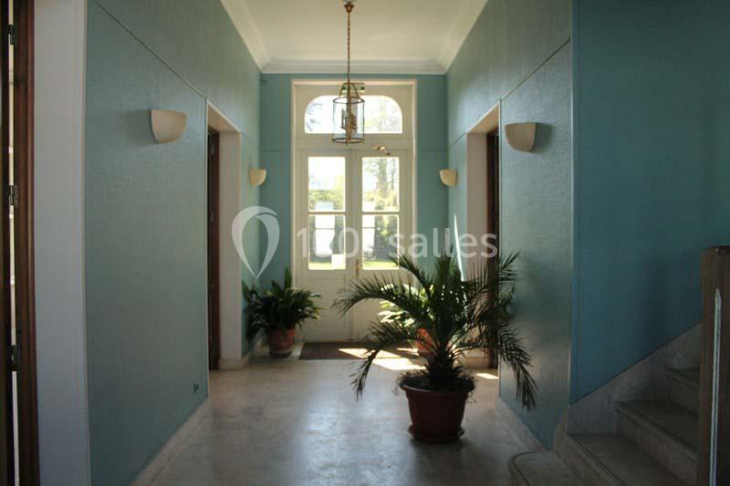 Couloir lumineux avec murs bleus, plantes en pot, escalier à droite et porte vitrée donnant sur un jardin.