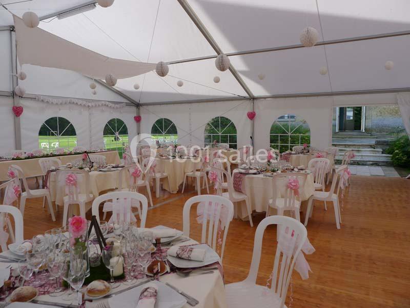 Salle de réception sous chapiteau avec tables décorées, nappes blanches, chaises en plastique et guirlandes suspendues.