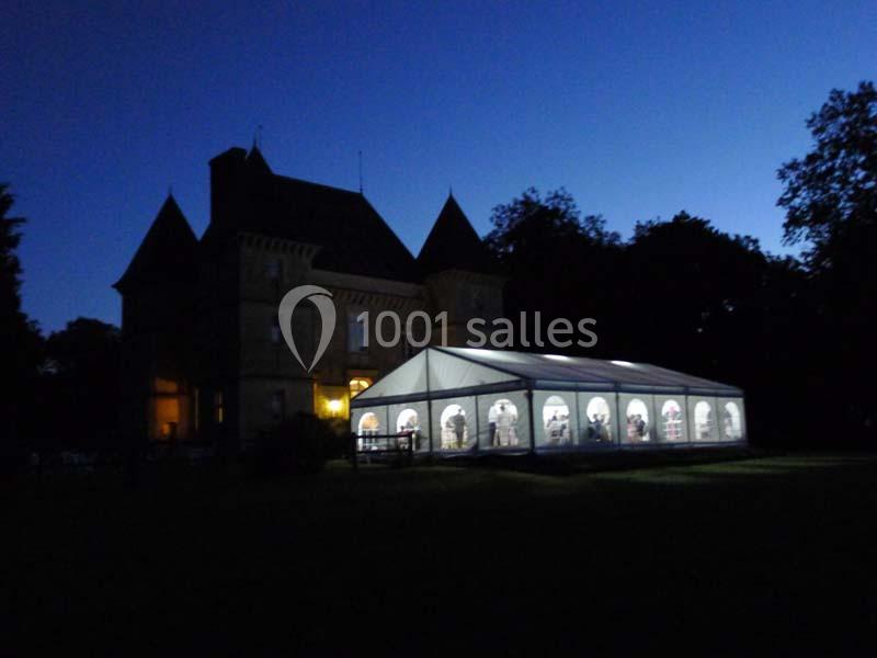 Grande tente éclairée installée devant un château au crépuscule, entourée d'arbres dans un cadre sombre.