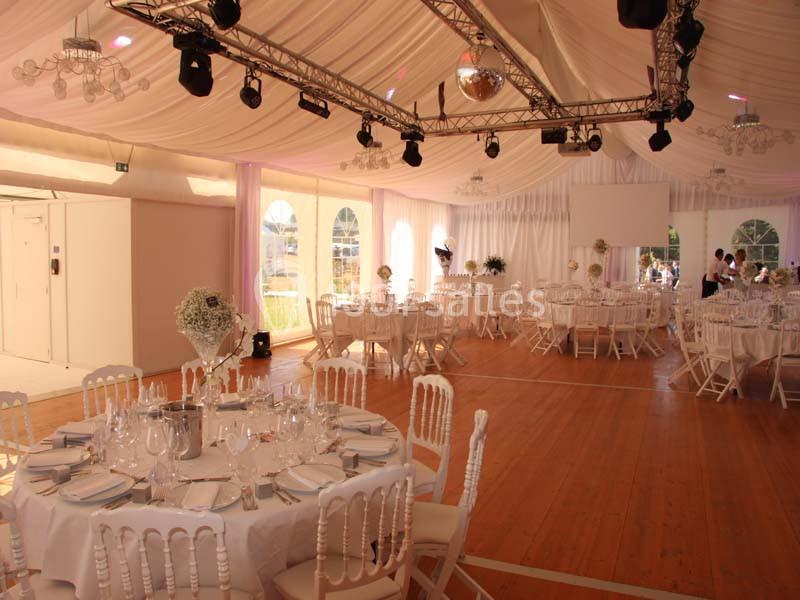 Salle de réception décorée avec des tables rondes dressées, chaises blanches et plafond drapé de tissu clair.