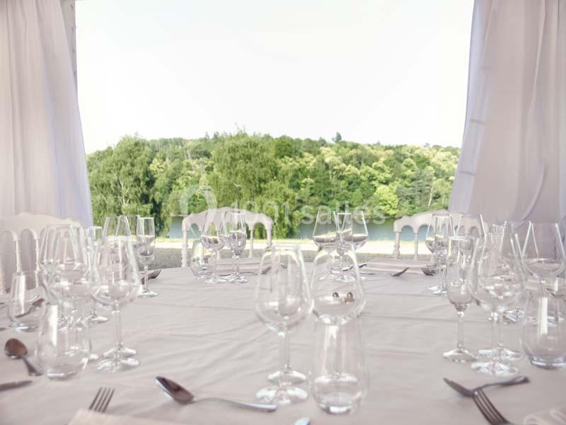 Table dressée avec nappes blanches, verres et couverts, offrant une vue sur un paysage verdoyant et un plan d'eau.