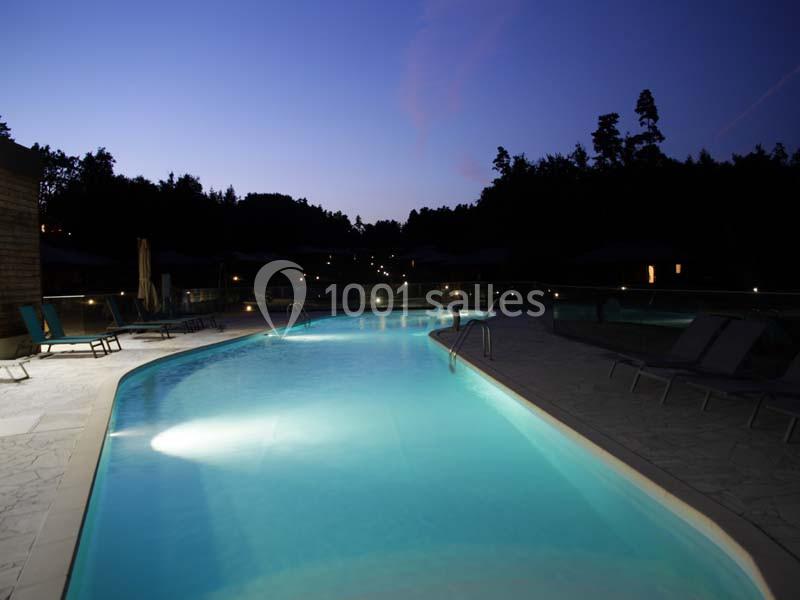 Piscine éclairée en soirée, entourée de chaises longues et bordée par des arbres dans un cadre paisible.