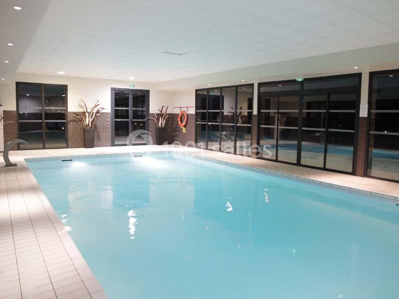 Piscine intérieure éclairée avec carrelage clair, grandes baies vitrées et plantes décoratives.