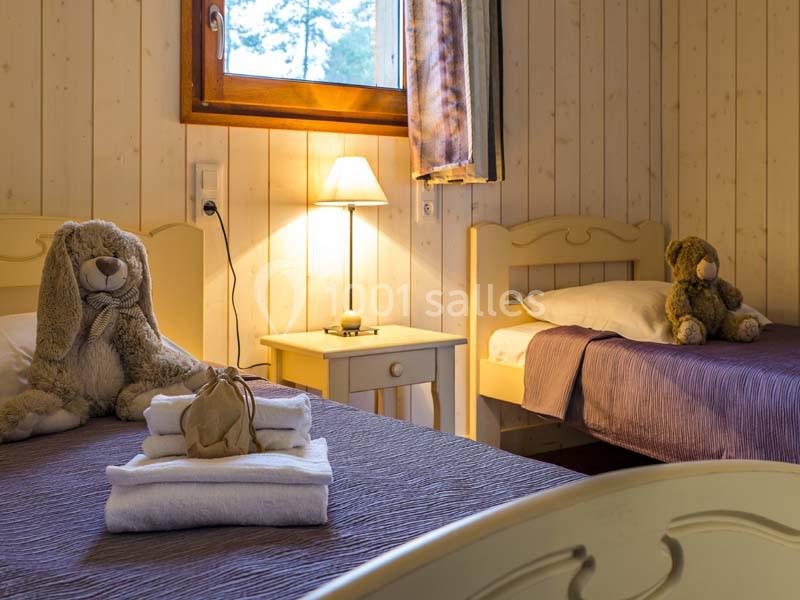 Deux lits simples avec linge de lit violet, une lampe allumée sur une table de chevet et des peluches posées sur les lits.