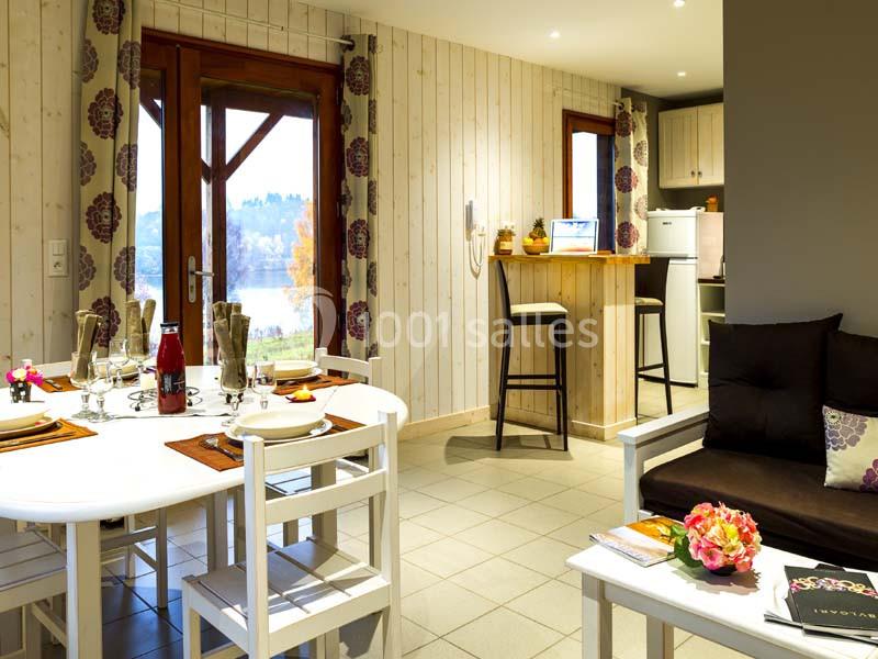 Salle à manger lumineuse avec table dressée, coin salon, cuisine ouverte et vue sur l'extérieur par une porte vitrée.