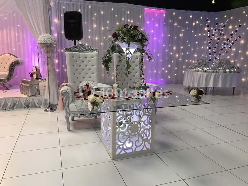 Salle de réception décorée avec des fauteuils élégants, une table en verre illuminée et des arrangements floraux.