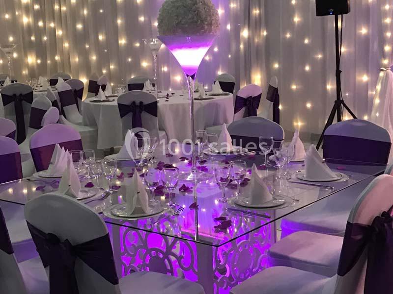 Salle de réception décorée avec des tables élégantes, nappes blanches, chaises ornées de rubans violets et éclairage tamisé.