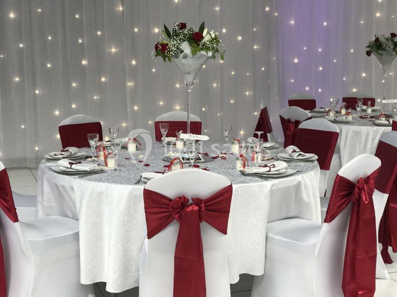 Salle décorée pour un événement, avec tables rondes dressées, nappes blanches, chaises ornées de nœuds rouges et éclairage…