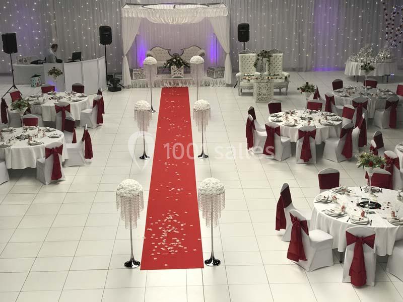 Salle de réception décorée avec des tables rondes, nappes blanches, chaises rouges et une allée centrale rouge ornée de…