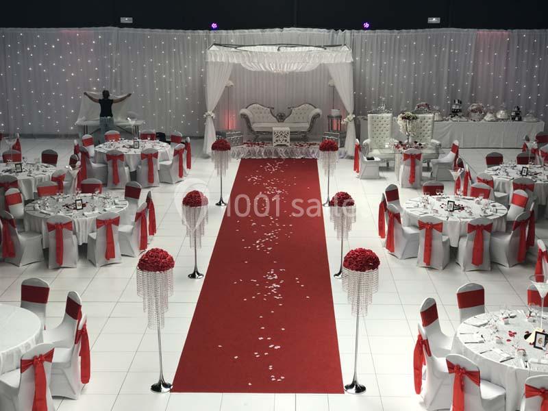 Salle de réception décorée avec des tables rondes, des chaises ornées de rubans rouges et une allée centrale rouge.