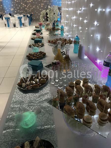 Buffet de réception avec verrines, petits sandwichs et boissons, disposé sur une table décorée de lumières et de motifs…