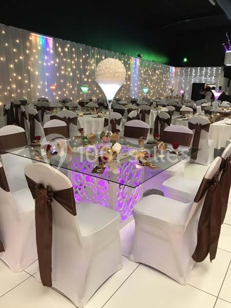 Salle de réception décorée avec des tables en verre, chaises blanches ornées de nœuds marron et éclairages lumineux.