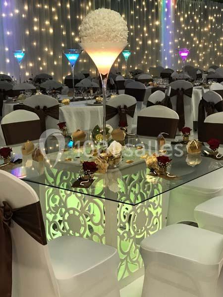 Salle de réception décorée avec des tables en verre illuminées, chaises blanches et centres de table élégants.
