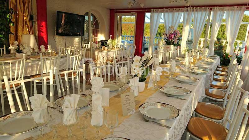 Salle de réception lumineuse avec tables décorées de nappes blanches, vaisselle élégante et serviettes pliées.