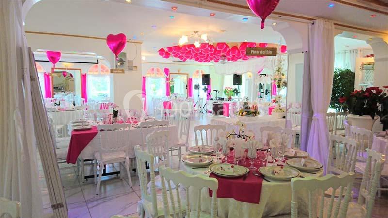 Salle de réception décorée avec des ballons et nappes roses, tables dressées avec vaisselle élégante.