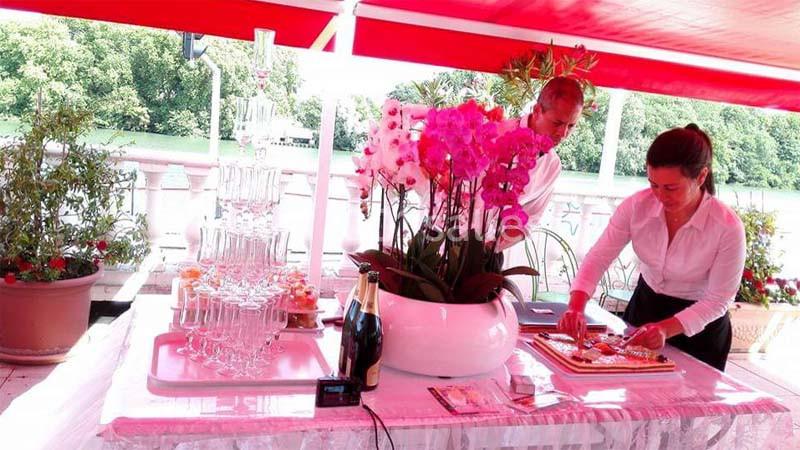 Deux personnes préparent un buffet sous un auvent rouge, avec des fleurs, des verres et des bouteilles de champagne.