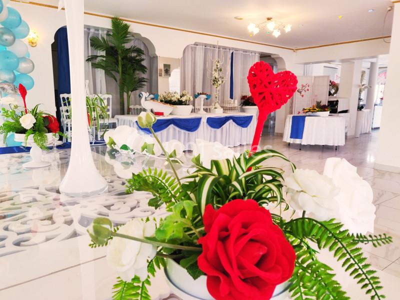 Salle décorée pour un événement avec des fleurs rouges et blanches au premier plan et des tables ornées en arrière-plan.