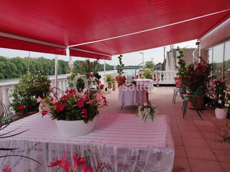 Terrasse couverte avec des tables décorées de nappes blanches et de fleurs, offrant une vue sur un fleuve et des arbres.