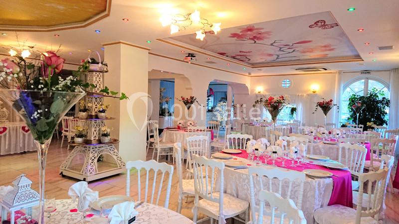 Salle de réception lumineuse décorée avec des tables rondes, nappes blanches, chemins de table roses et arrangements floraux.
