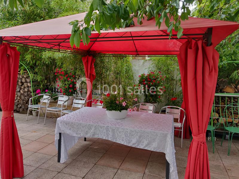 Tonnelle rouge avec rideaux, table recouverte d'une nappe blanche et pot de fleurs, entourée de chaises et de végétation.